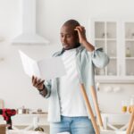 Confused black man having hard time assembling furniture and reading self-assembly manual in kitchen