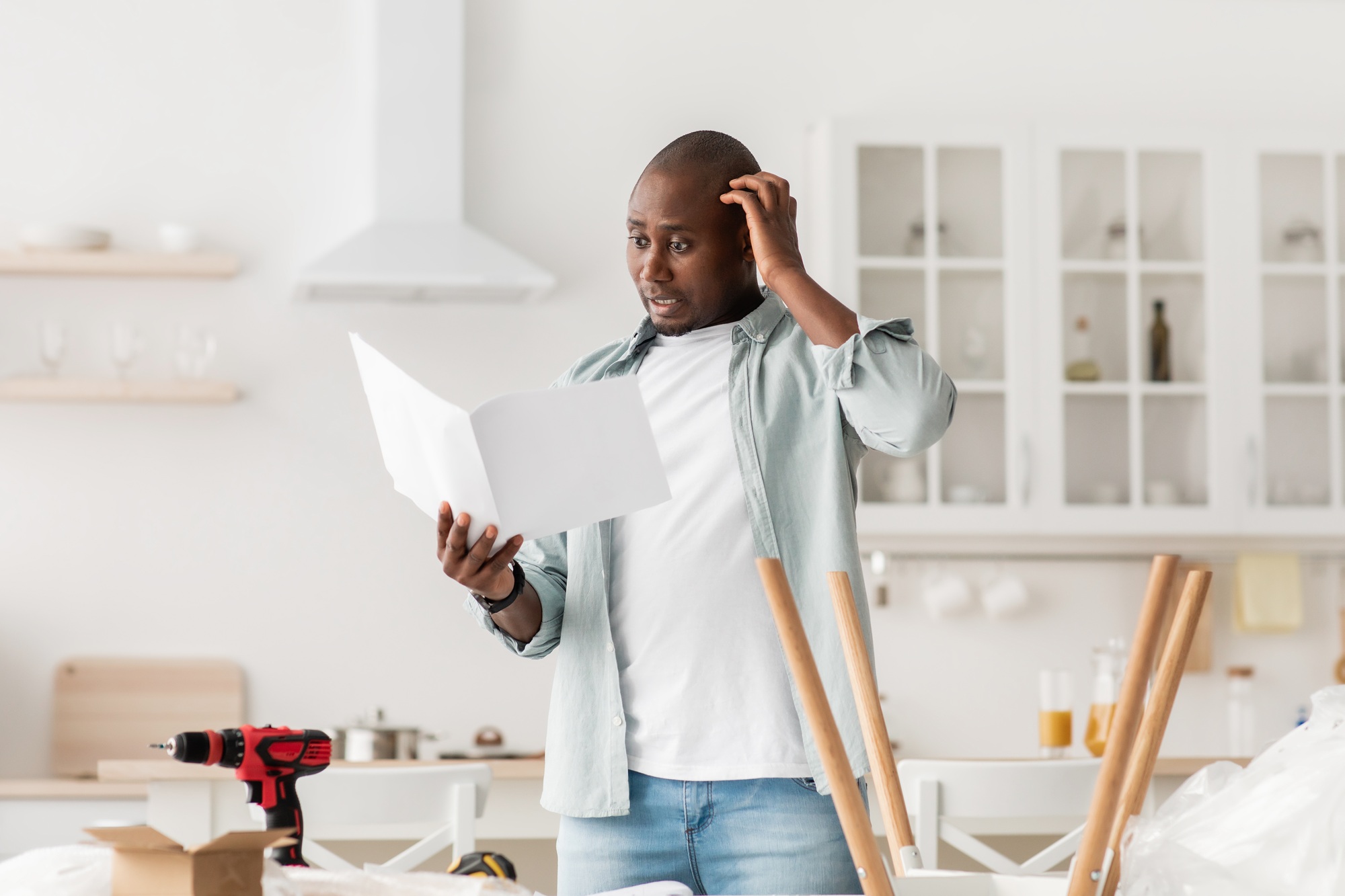 Confused black man having hard time assembling furniture and reading self-assembly manual in kitchen